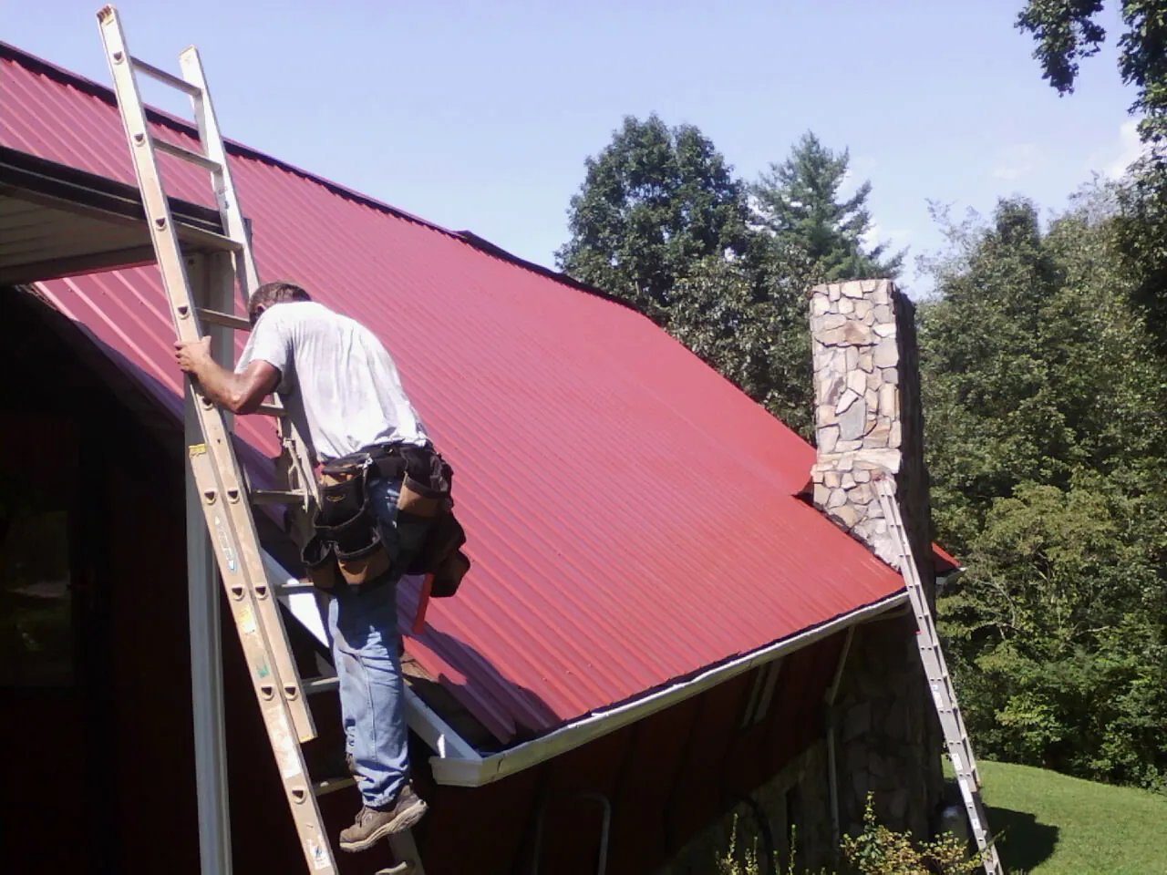Roofer on a ladder beside a red metal roof and stone chimney — Soffit & Fascia Repair by Roofing Brothers in Sumiton